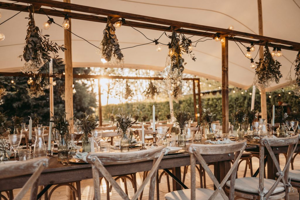 Sfeervolle buitenbruiloft onder een tent met lange houten tafels, prachtig gedekt met glaswerk, kaarsen en wilde bloemen in vazen. Houten stoelen met kruisvormige rugleuningen omringen de tafels. Boven de tafels hangen sfeerverlichting en gedroogde bloemen, terwijl de zonsondergang een warme, romantische gloed over de setting werpt