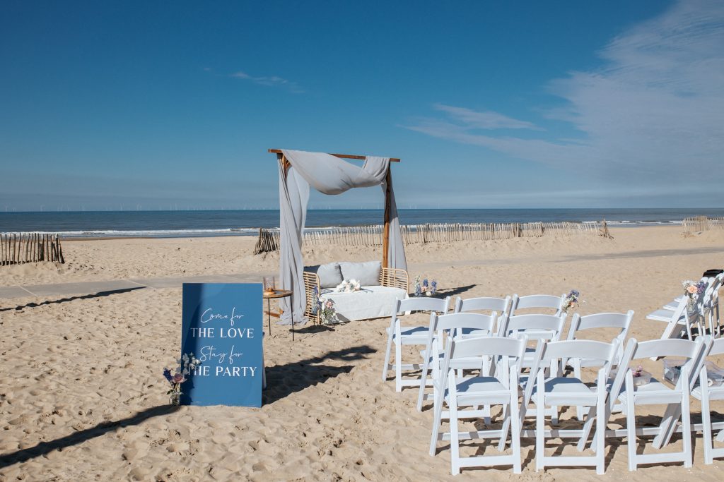 Romantische strandbruiloftsopstelling met een houten prieel versierd met lichte doeken en bloemen, witte stoelen gerangschikt voor de ceremonie en een blauw bord met de tekst: 'Come for the love, stay for the party'. Op de achtergrond een prachtig uitzicht op de zee en een heldere blauwe lucht.