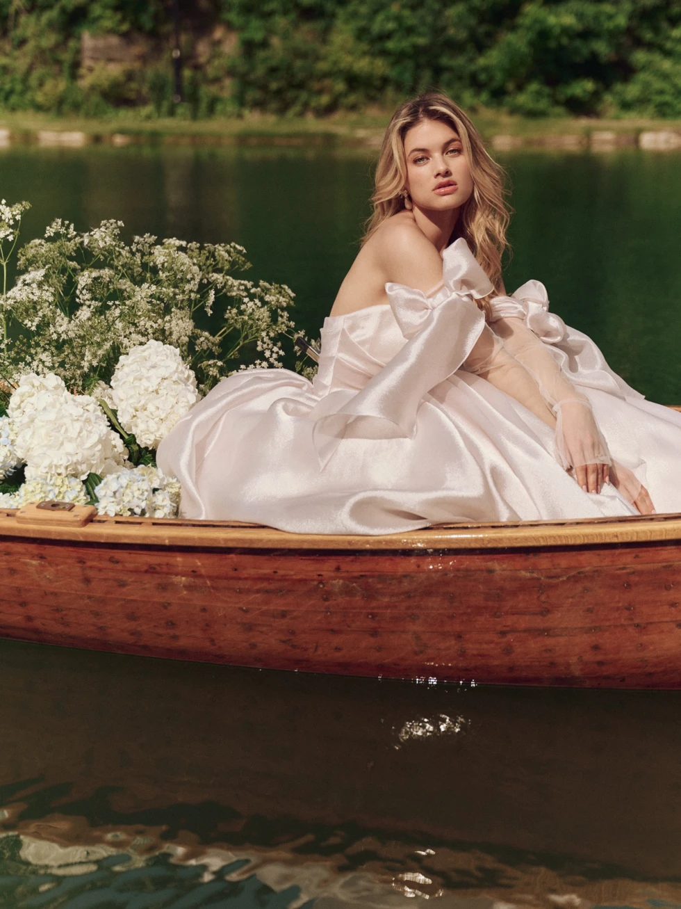 Een bruid in een elegante, off-the-shoulder baljurk met grote strikdetails en een glanzende stof zit dromerig in een houten boot op een rustig meer. De boot is versierd met witte bloemen en groene takken, terwijl het water en het groene landschap een idyllische sfeer creëren.