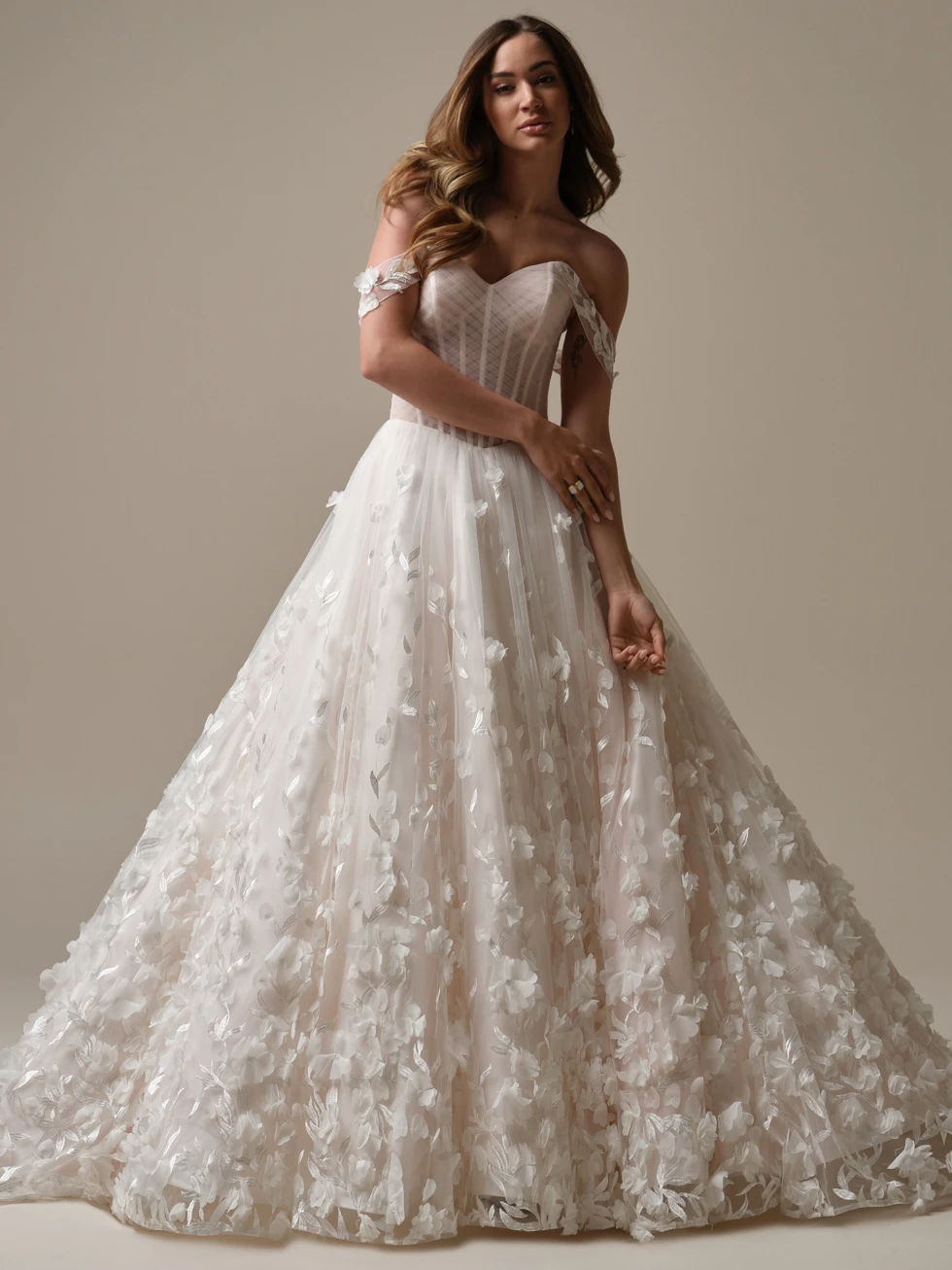 Een bruid draagt een romantische baljurk trouwjurk met een doorzichtig korset en off-the-shoulder bandjes versierd met delicate bloemenapplicaties. De volumineuze tule rok is rijkelijk versierd met 3D-bloemen en zilveren bladaccenten, wat een sprookjesachtige uitstraling creëert. Haar lange, golvende haar en elegante houding versterken de dromerige sfeer van de look.