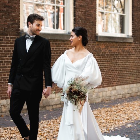 Een stralend bruidspaar loopt hand in hand over een met herfstbladeren bedekte straat, omringd door historische bakstenen gebouwen met grote ramen. De bruid draagt een elegante witte jurk met volumineuze mouwen en een lange sleep, terwijl ze een boeket met droogbloemen vasthoudt. De bruidegom draagt een stijlvol zwart pak met een vlinderdas. Ze kijken elkaar verliefd aan.