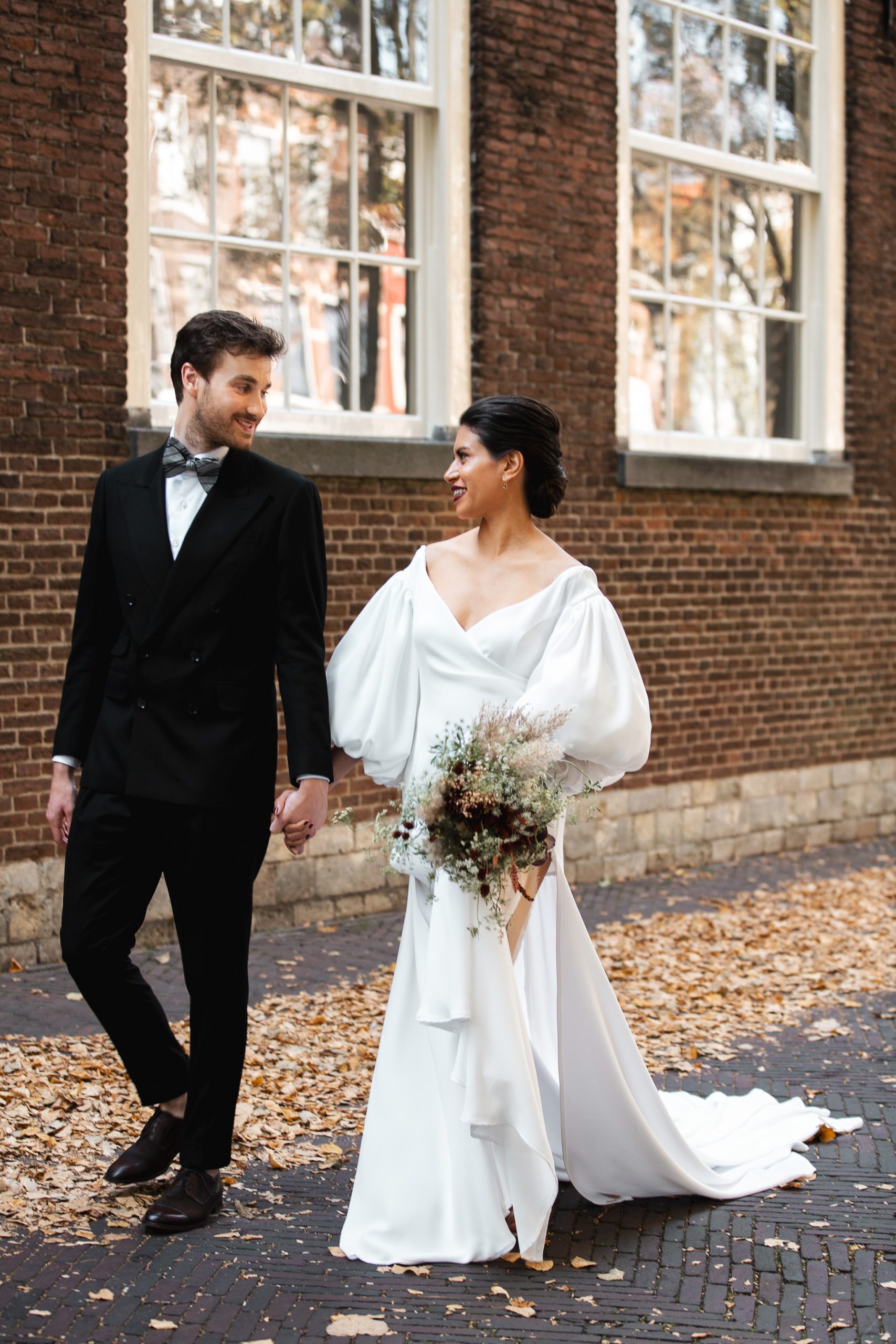 Een stralend bruidspaar loopt hand in hand over een met herfstbladeren bedekte straat, omringd door historische bakstenen gebouwen met grote ramen. De bruid draagt een elegante witte jurk met volumineuze mouwen en een lange sleep, terwijl ze een boeket met droogbloemen vasthoudt. De bruidegom draagt een stijlvol zwart pak met een vlinderdas. Ze kijken elkaar verliefd aan.