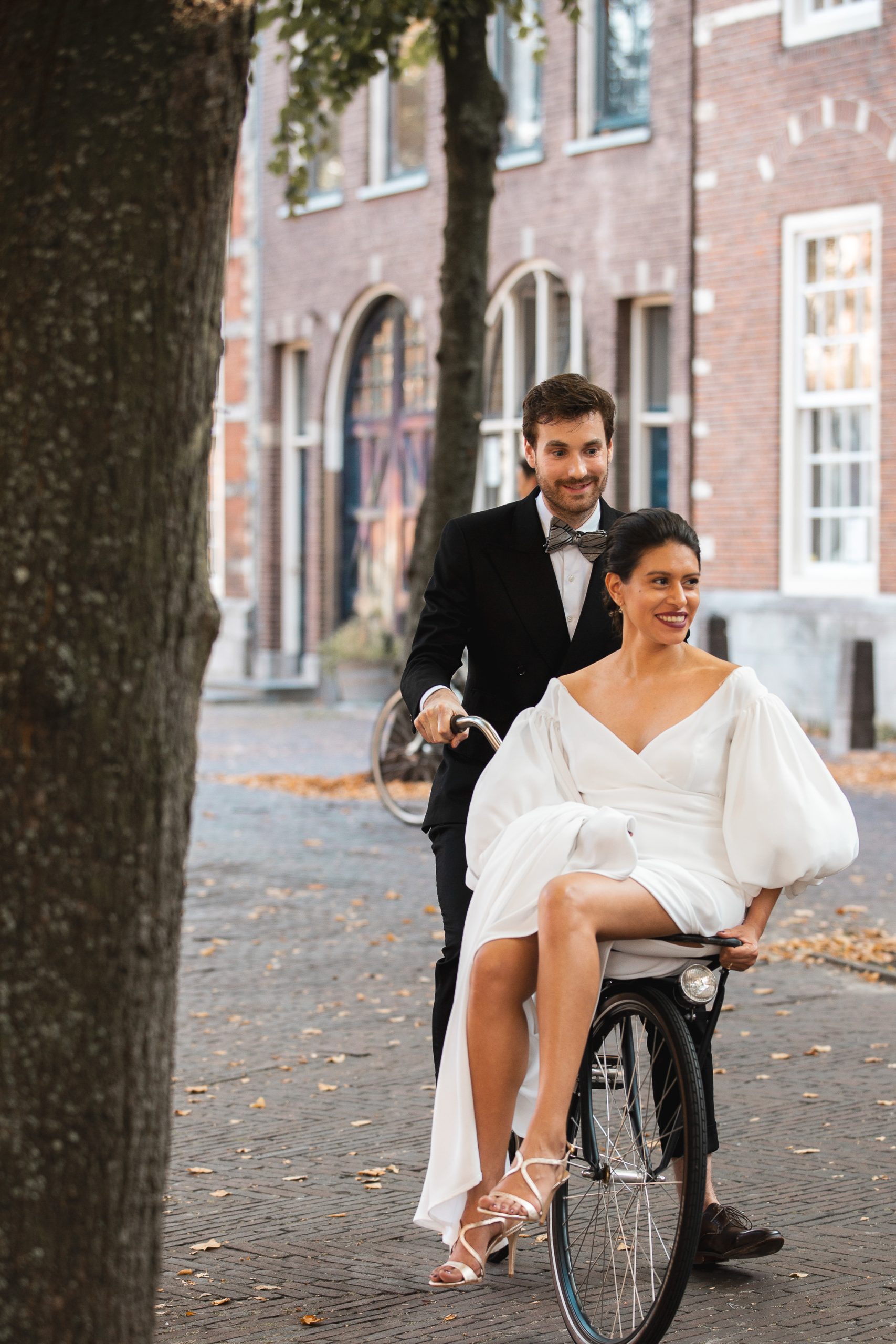 Een bruid en bruidegom genieten van een speels moment op hun trouwdag. De bruid, gekleed in een elegante witte jurk met volumineuze mouwen, zit lachend op het stuur van een vintage fiets, terwijl de bruidegom in een klassiek zwart pak haar voortduwt. De scène speelt zich af in een sfeervolle, historische straat met bakstenen gebouwen en herfstbladeren op de grond.