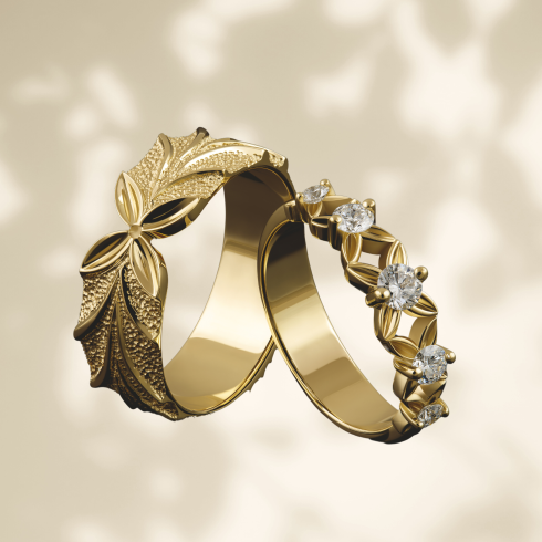 Een set elegante gouden trouwringen met een verfijnd natuurmotief. De ene ring heeft een gedetailleerd bladpatroon met een gestructureerde afwerking, terwijl de andere is versierd met fonkelende diamanten en delicate bladvormige zettingen. De ringen zweven sierlijk tegen een zachte, neutrale achtergrond met schaduwpatronen.