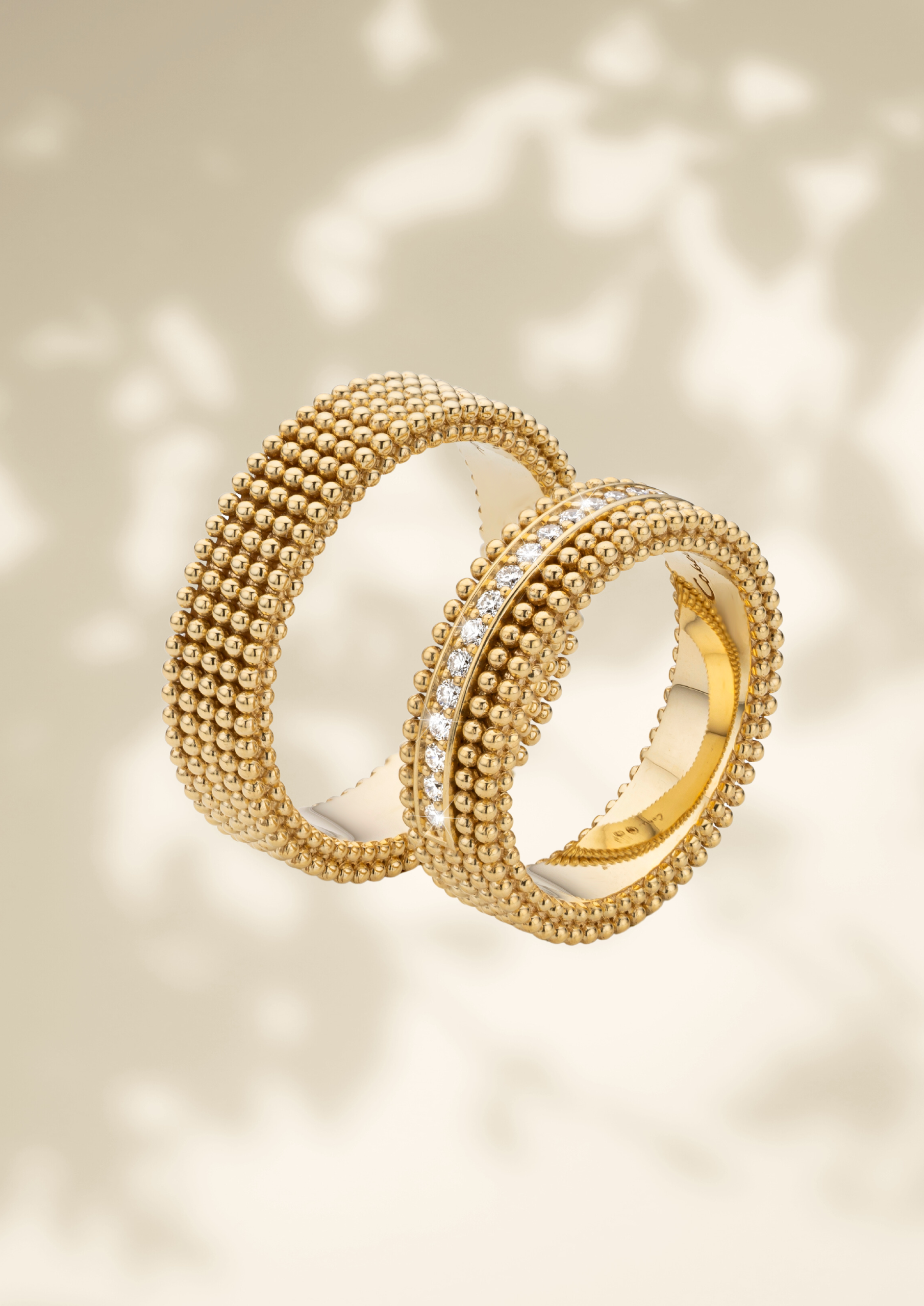 Een set exclusieve gouden trouwringen met een verfijnde bolletjesstructuur. Eén ring heeft een volledig gedetailleerd ontwerp, terwijl de andere is verrijkt met een rij sprankelende diamanten in het midden. De ringen zweven elegant tegen een zachte, neutrale achtergrond met subtiele licht- en schaduweffecten.