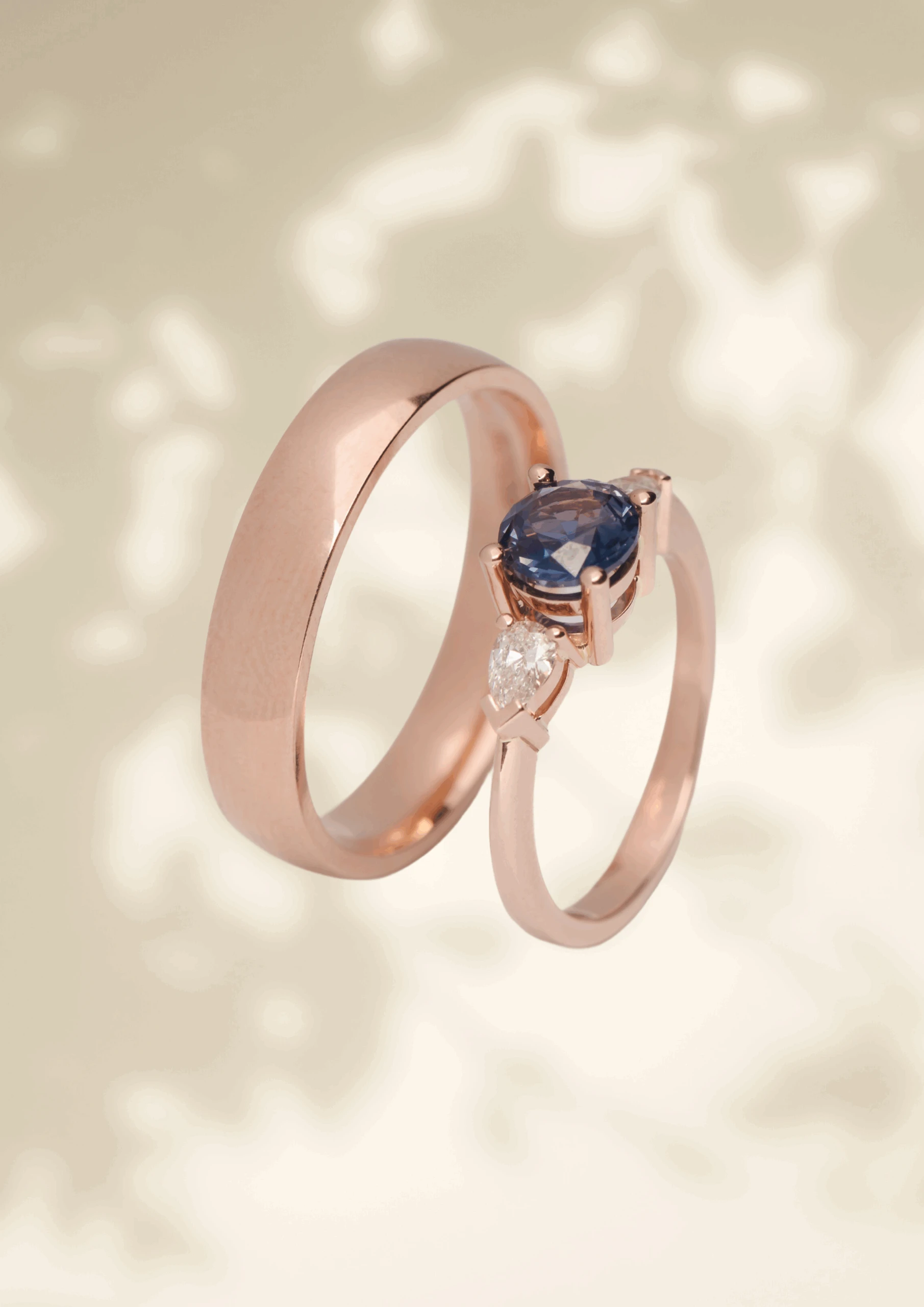 Een elegante set roségouden trouwringen. De ene ring is een klassieke, gladde trouwring, terwijl de andere is voorzien van een schitterende blauwe edelsteen in een verfijnde zetting, aangevuld met een peervormige diamant. De ringen zweven tegen een zachte, neutrale achtergrond met subtiele licht- en schaduweffecten.
