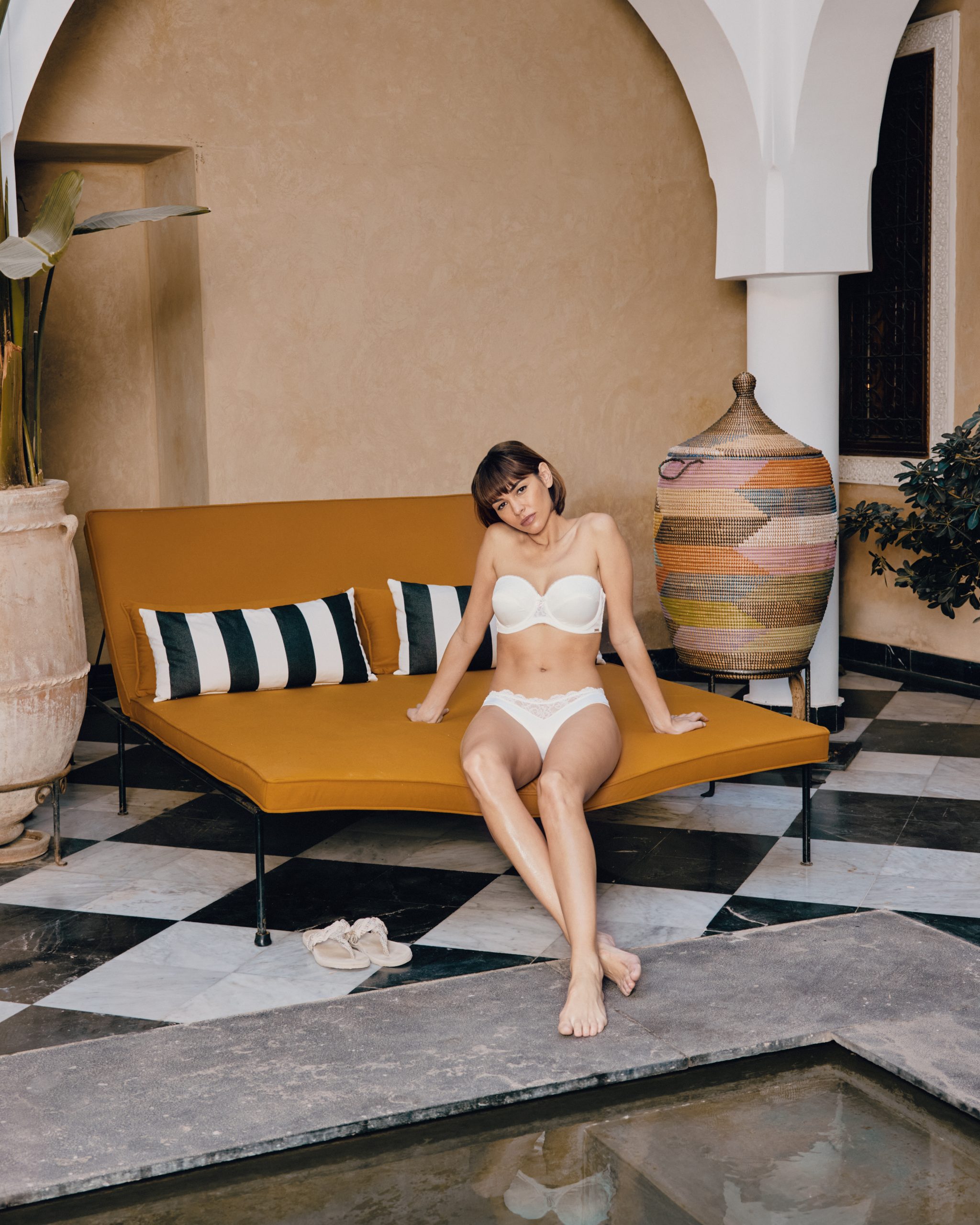Een vrouw in elegante witte lingerie uit de 'Pure Love' collectie zit ontspannen op een okergele loungebank in een sfeervolle patio met Marokkaanse invloeden. De ruimte heeft zwart-wit geblokte vloertegels, aardetinten op de muren, en decoratieve elementen zoals een kleurrijke mand en tropische planten. Op de voorgrond is een klein waterbassin zichtbaar, wat een serene sfeer toevoegt.