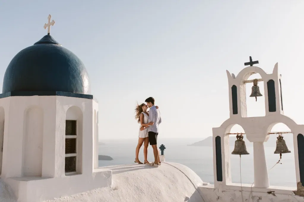 Een verliefd koppel deelt een intiem moment op het witte dak van een iconische Griekse kerk in Santorini, omringd door een adembenemend uitzicht op de zee. De blauwe koepel en de klokkentoren met belletjes omlijsten het tafereel, terwijl de zon een warme gloed over de scène werpt. De wind speelt met de haren van de vrouw terwijl ze dicht bij elkaar staan en genieten van het moment.