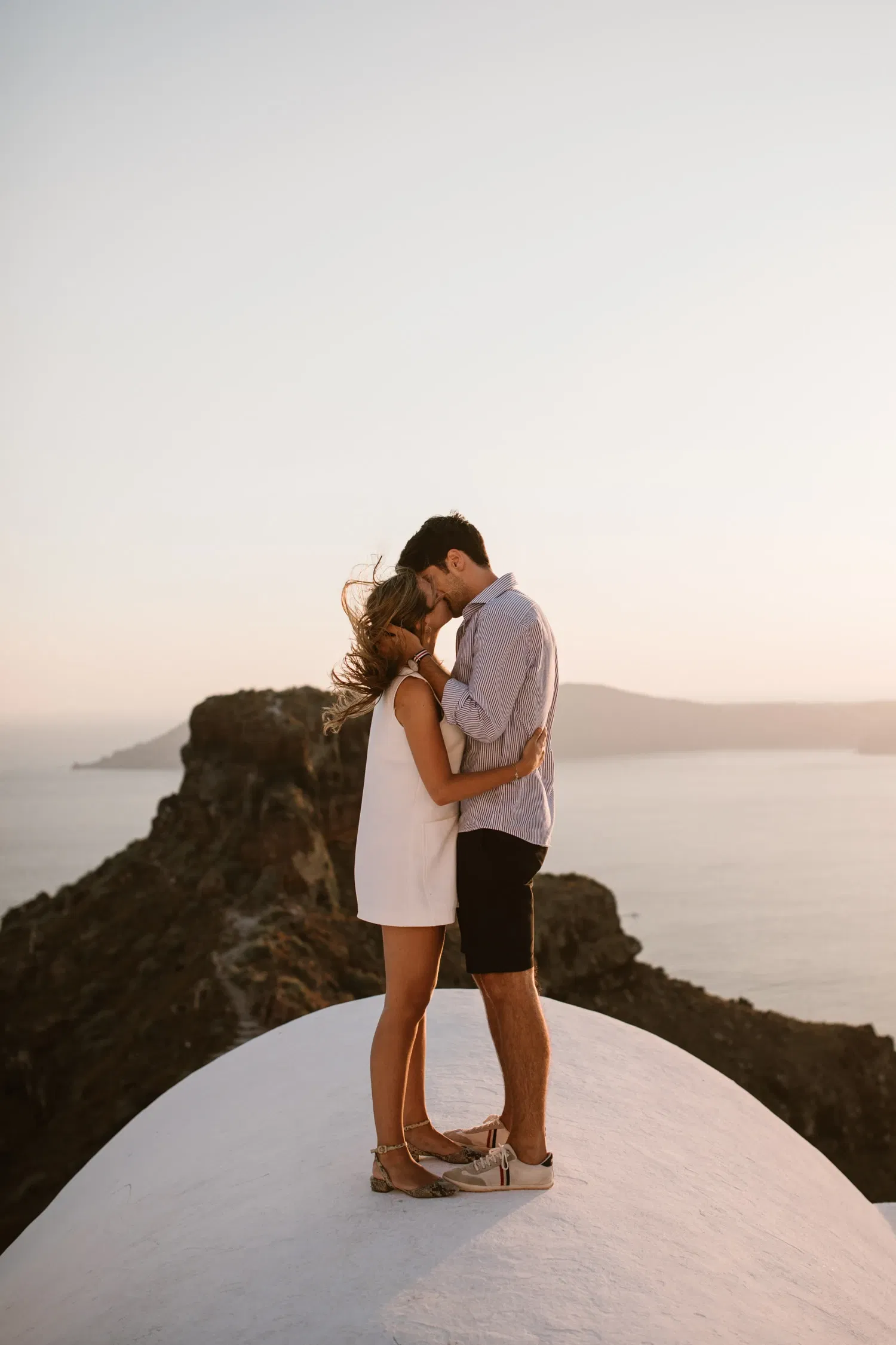 Een verliefd koppel staat op het witte dak van een gebouw in Santorini, omringd door een adembenemend uitzicht op de zee en ruige kliffen. De man houdt teder het gezicht van de vrouw vast terwijl hun voorhoofden elkaar raken. De warme gloed van de ondergaande zon verlicht hun silhouetten en de wind speelt met haar haren, wat een dromerige en romantische sfeer creëert.