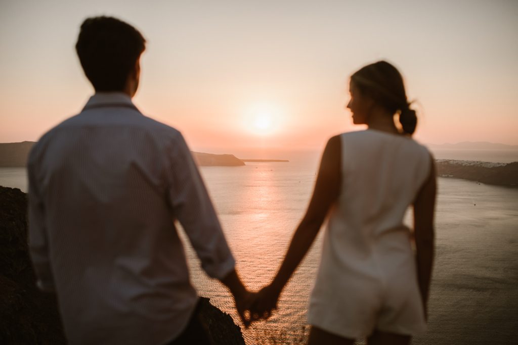 Een silhouet van een koppel dat hand in hand staat en samen geniet van een prachtige zonsondergang boven de zee. De warme gloed van de zon reflecteert op het water terwijl de horizon zich uitstrekt over de heuvelachtige kustlijn.