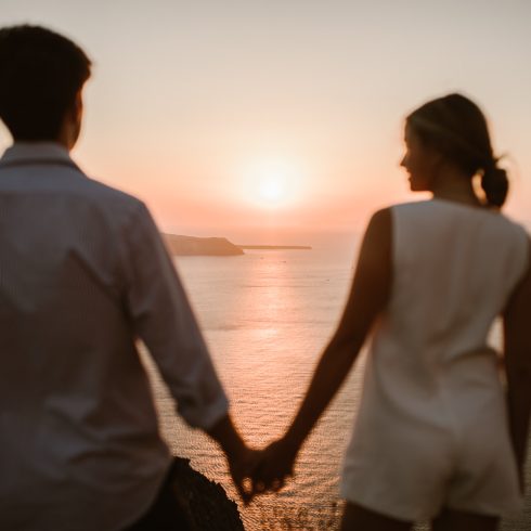 Een silhouet van een koppel dat hand in hand staat en samen geniet van een prachtige zonsondergang boven de zee. De warme gloed van de zon reflecteert op het water terwijl de horizon zich uitstrekt over de heuvelachtige kustlijn.