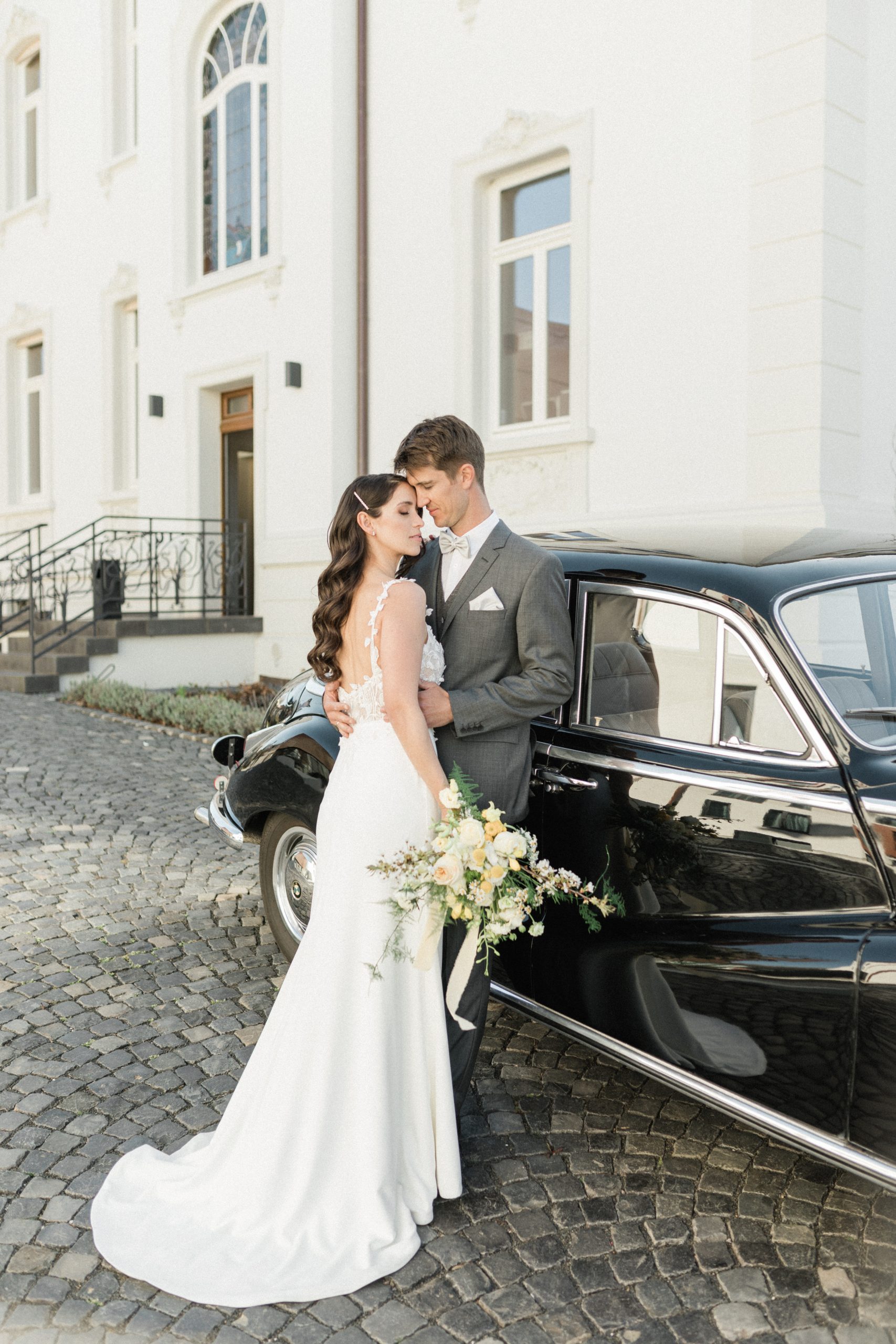 Bruid en bruidegom in een intiem moment naast een glanzende zwarte oldtimer, met op de achtergrond een wit historisch gebouw en geplaveide straat.