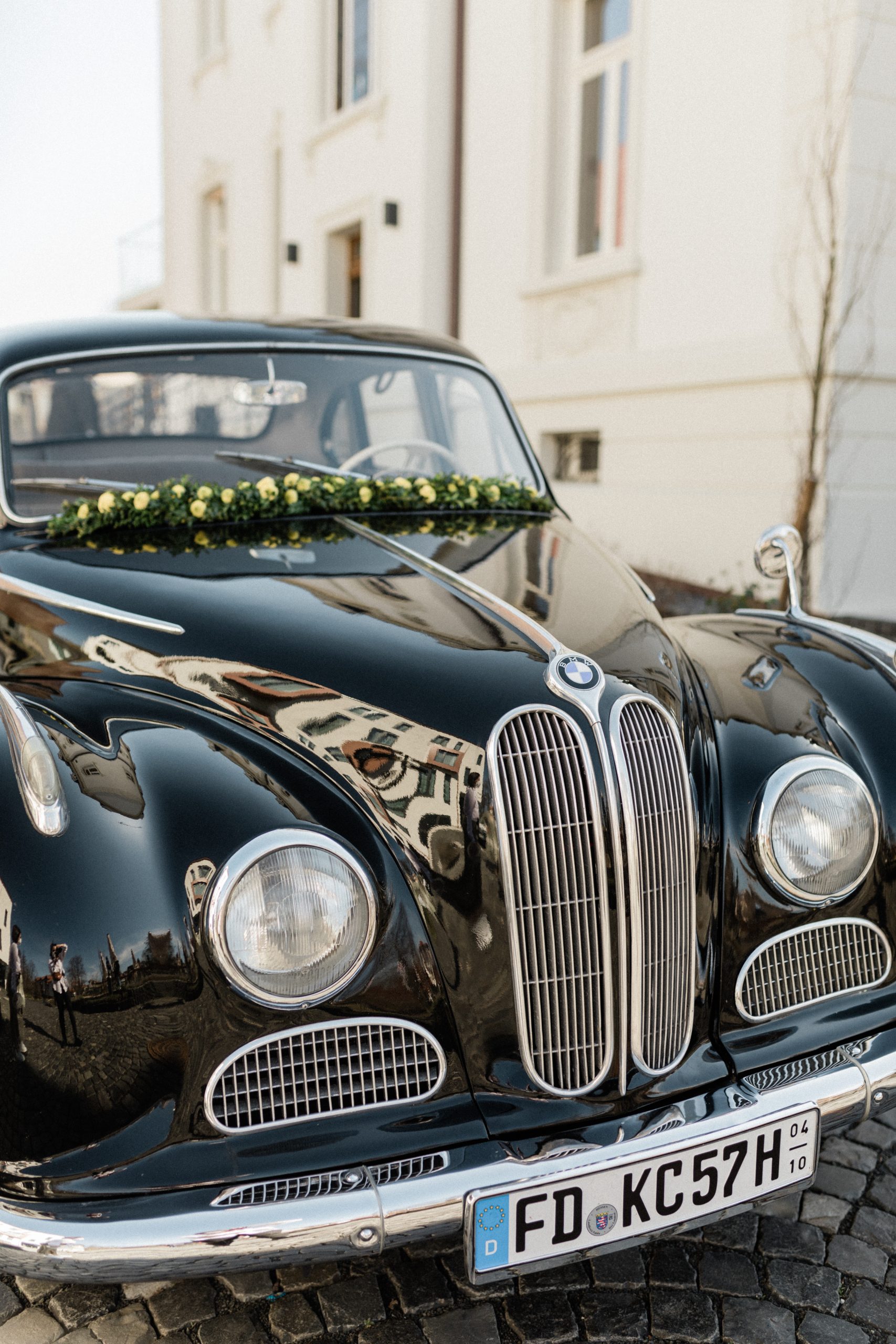 lanzende zwarte klassieke BMW versierd met een bloemenkrans van gele rozen op de motorkap, geparkeerd voor een elegant wit gebouw op een geplaveide straat.