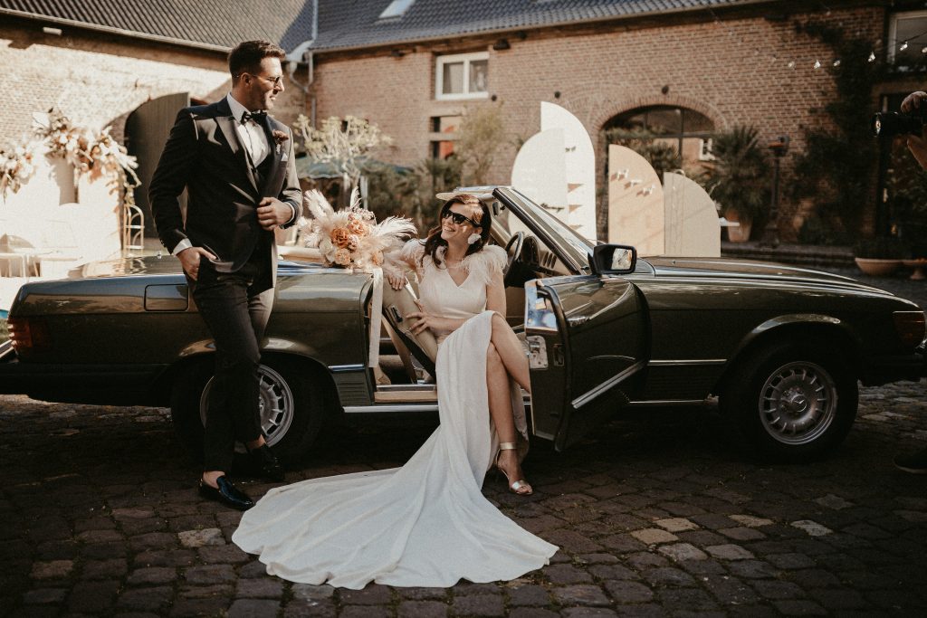 Bruid in een elegante witte jurk zit glimlachend in een klassieke cabriolet, terwijl de bruidegom in stijlvol pak naast de auto leunt, op een sfeervolle binnenplaats met romantische decoratie.