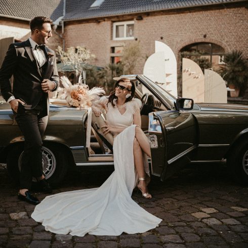Bruid in een elegante witte jurk zit glimlachend in een klassieke cabriolet, terwijl de bruidegom in stijlvol pak naast de auto leunt, op een sfeervolle binnenplaats met romantische decoratie.