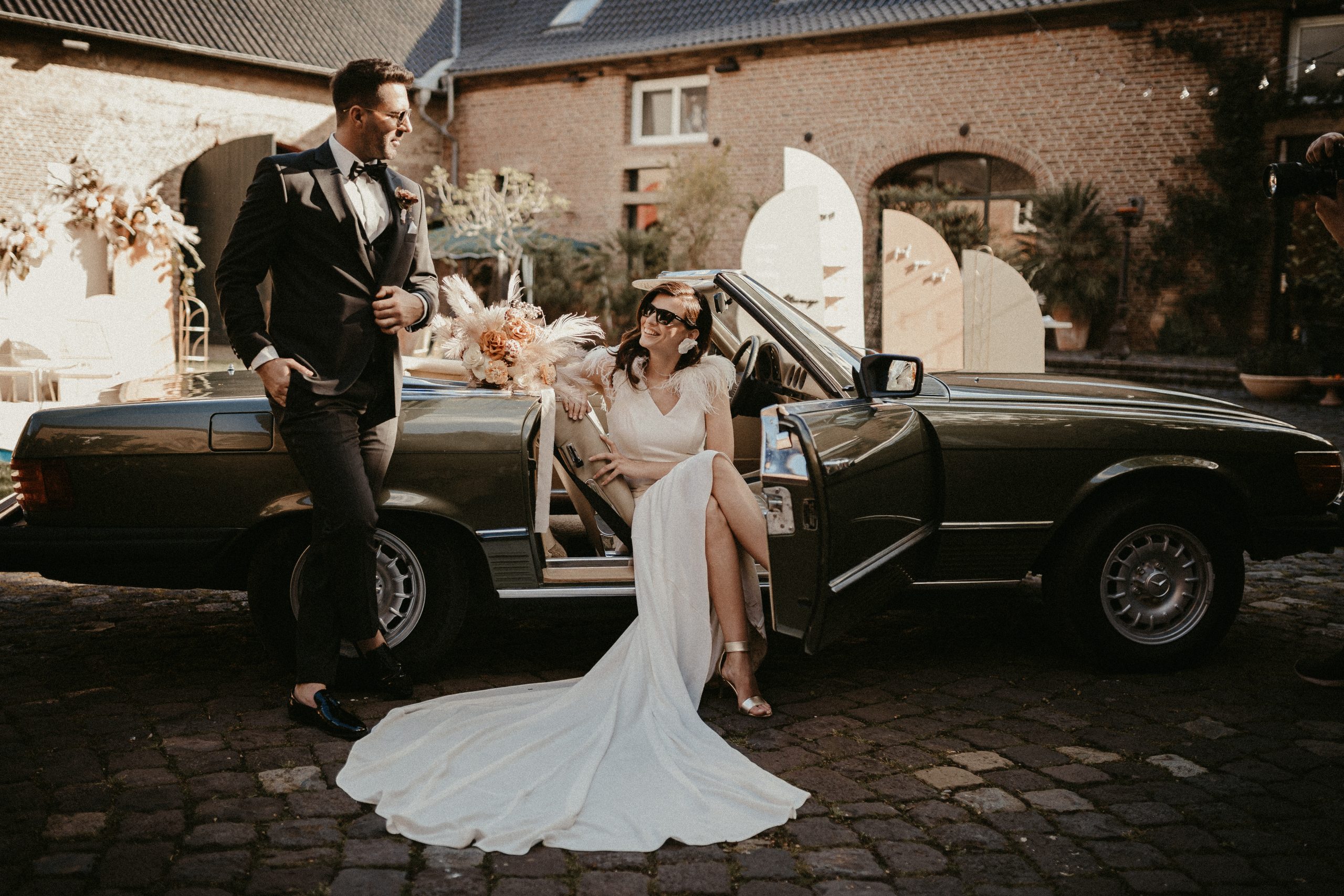 Bruid in een elegante witte jurk zit glimlachend in een klassieke cabriolet, terwijl de bruidegom in stijlvol pak naast de auto leunt, op een sfeervolle binnenplaats met romantische decoratie.