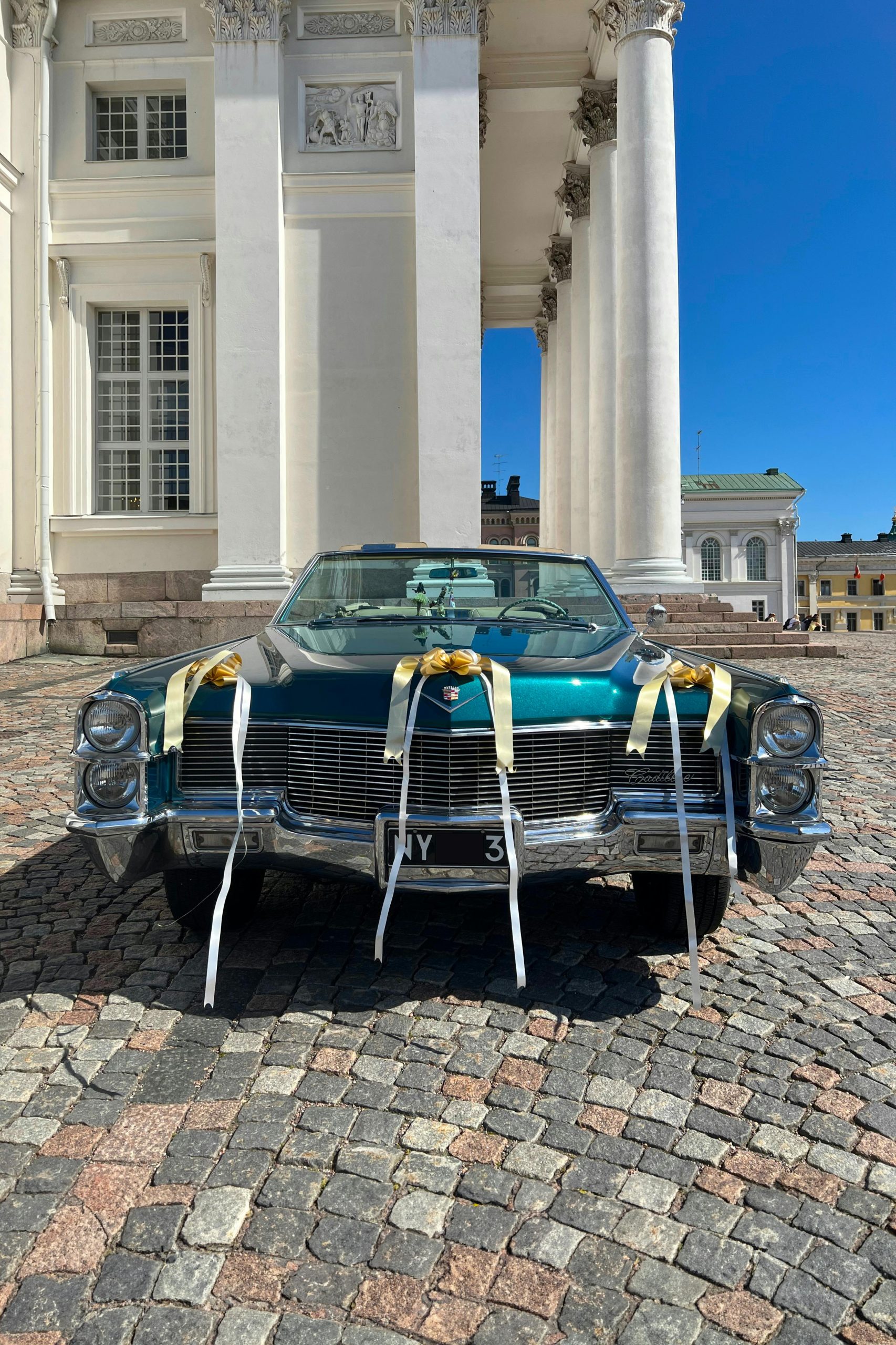 Versierde turquoise Cadillac oldtimer met witte en gouden linten, geparkeerd op een plein voor een klassiek wit gebouw met zuilen en heldere blauwe lucht.