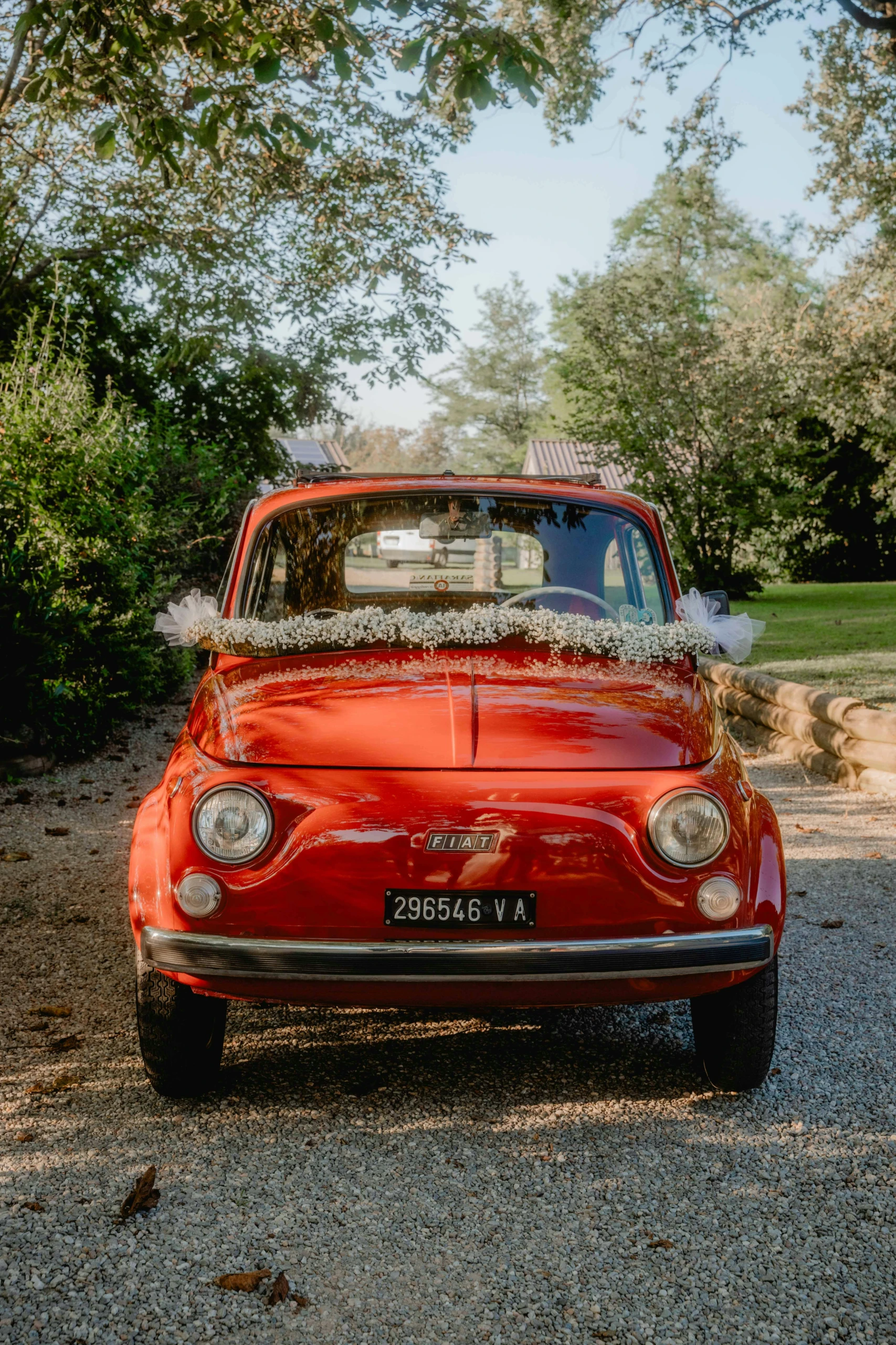 Vintage rode Fiat 500, versierd met bloemen en tule, staat geparkeerd op een landweggetje omringd door groen, klaar voor een bruiloft.