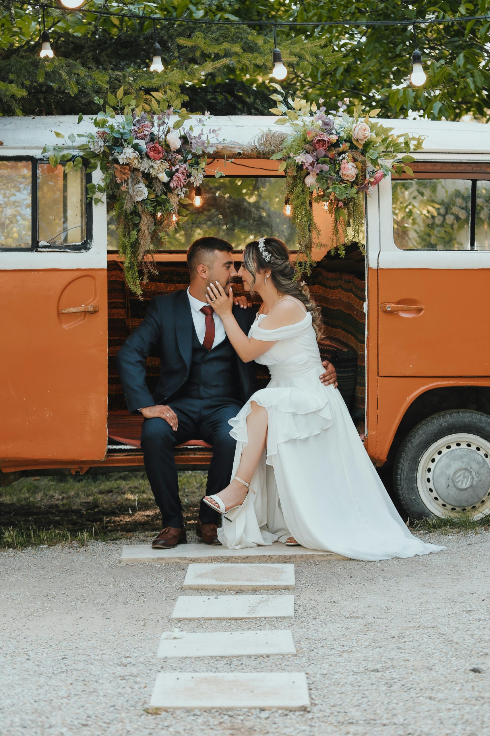 Bruid en bruidegom delen een teder moment zittend in een versierde oranje vintagebus, omringd door bloemen en lichtjes in een groene buitenomgeving.