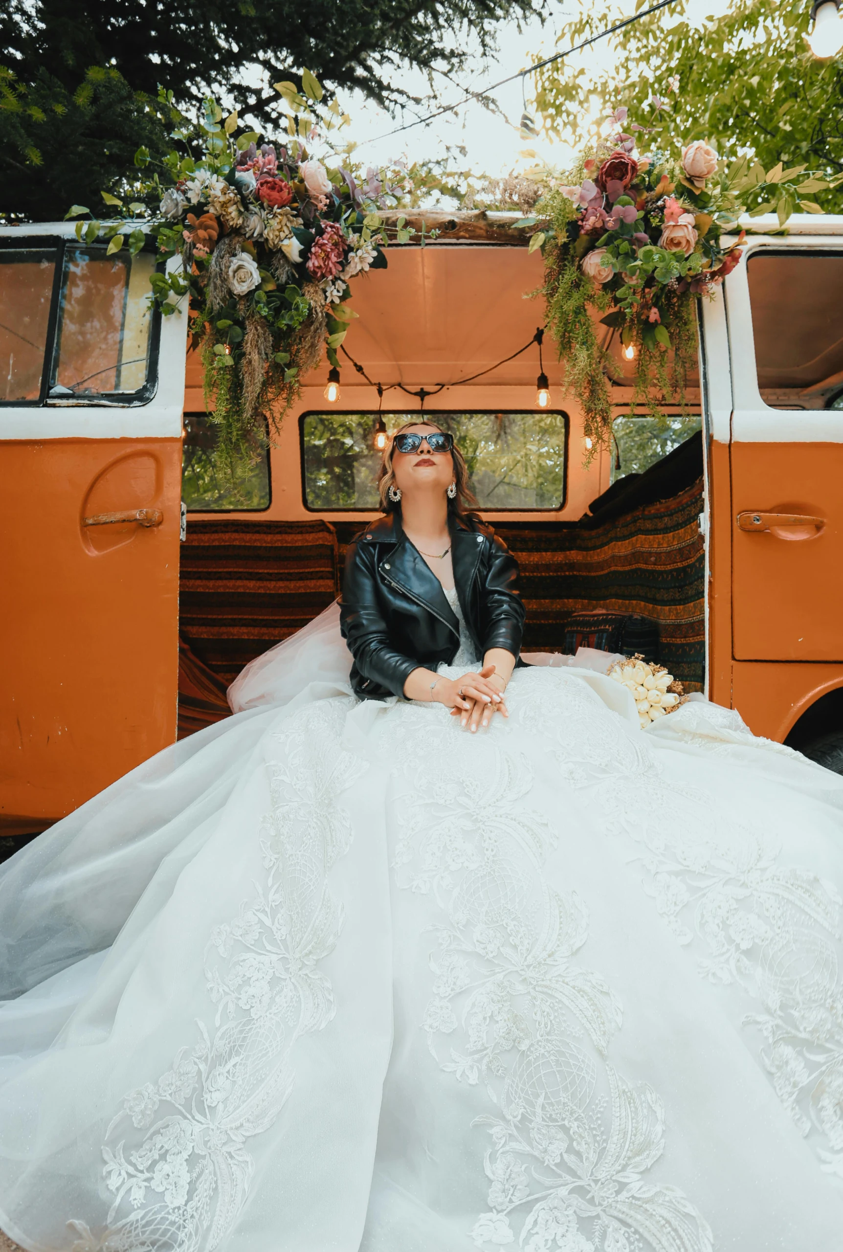 Bruid in een opvallende witte trouwjurk en leren jasje zit ontspannen voor een oranje retrobus, omringd door bloemen en lichtslingers, met een zelfverzekerde blik en zonnebril.