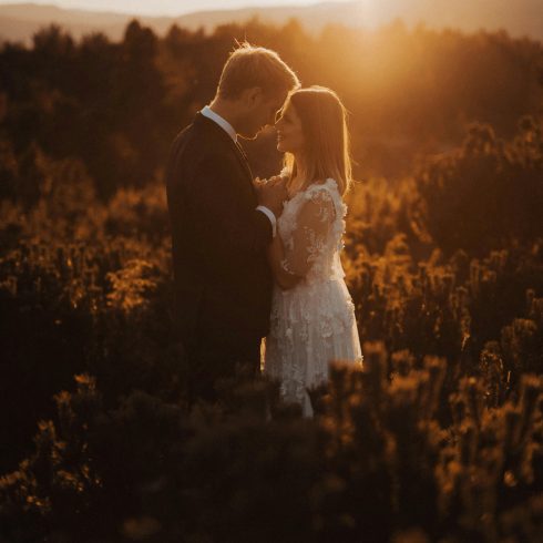 Bruid en bruidegom staan dicht bij elkaar in een veld tijdens zonsondergang, met warm gouden licht dat hun silhouetten omhult in een romantische, dromerige sfeer.