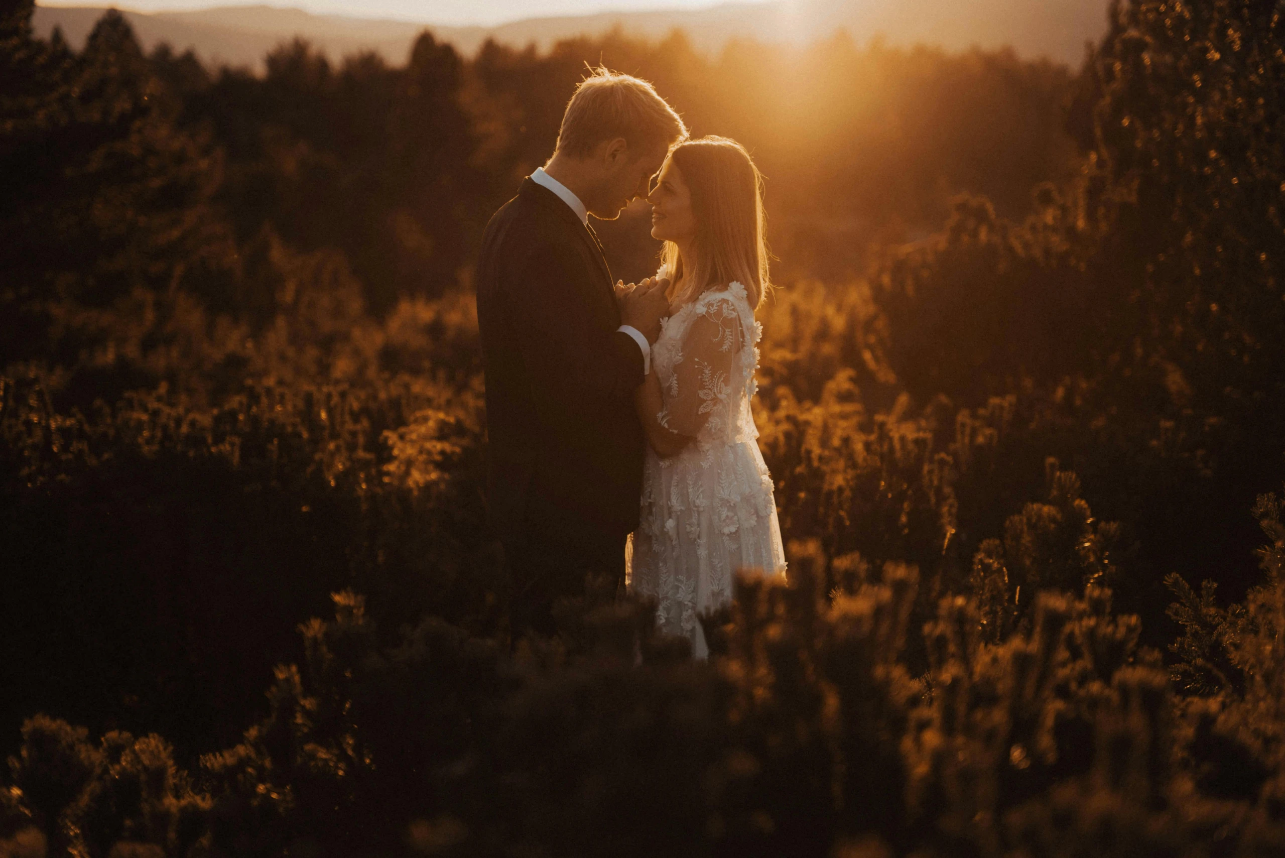 Bruid en bruidegom staan dicht bij elkaar in een veld tijdens zonsondergang, met warm gouden licht dat hun silhouetten omhult in een romantische, dromerige sfeer.