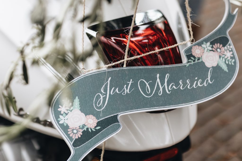 Close-up van een ‘Just Married’-bordje met bloemenprint, bevestigd aan de achterkant van een wit voertuig, met decoratieve takjes op de achtergrond.