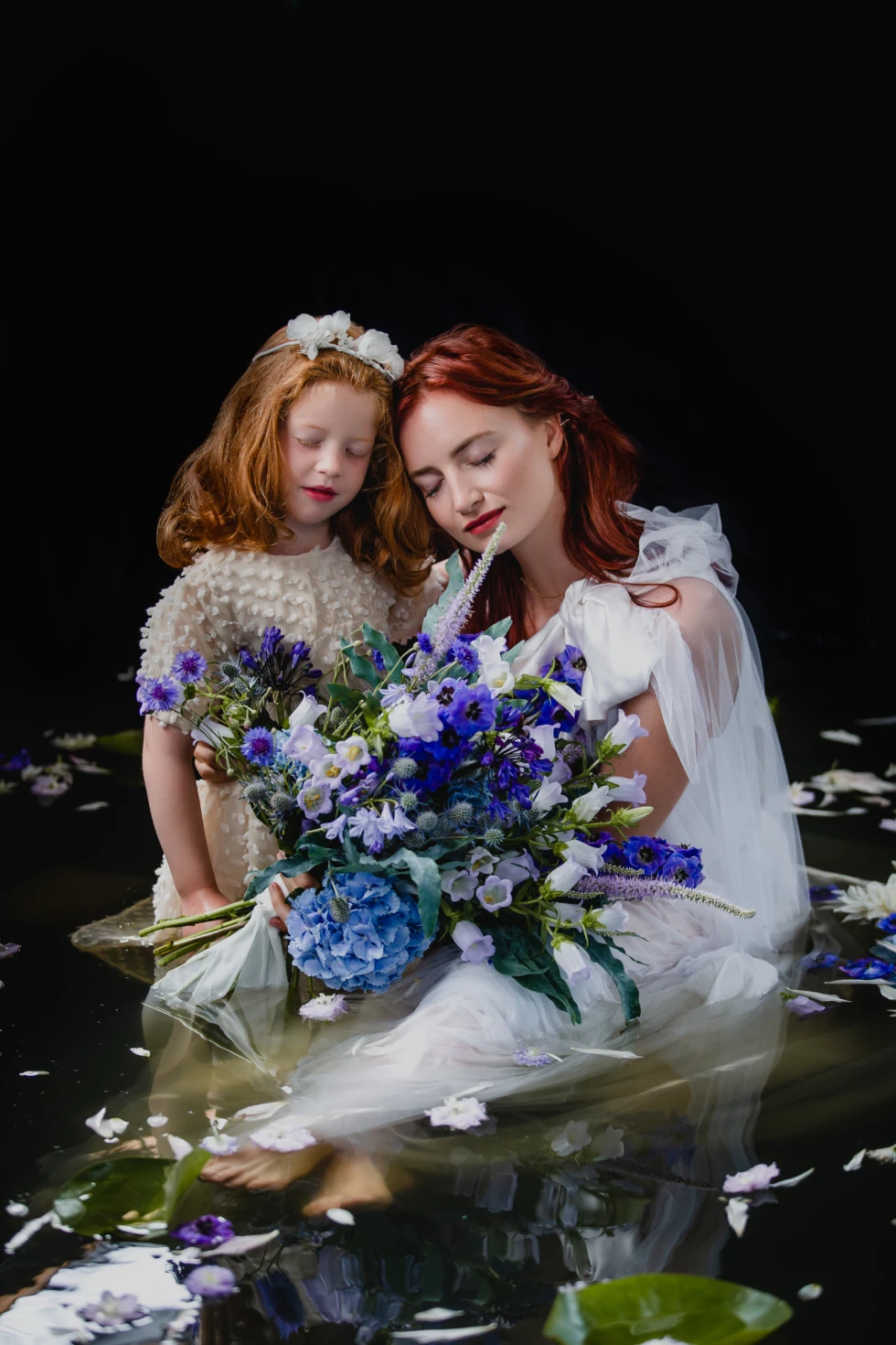 Sfeervol, sprookjesachtig portret van een vrouw en een jong meisje met gesloten ogen, omgeven door bloemen en water, in zachte belichting tegen een donkere achtergrond, waardoor een dromerige en magische sfeer ontstaat.