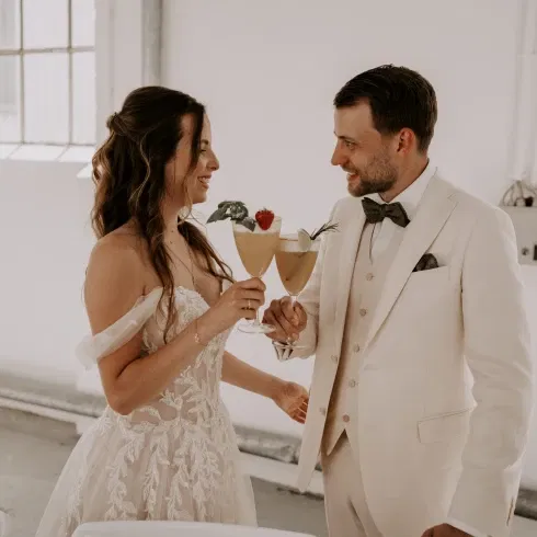 Bruid en bruidegom proosten met cocktails tijdens hun bruiloft, beiden gekleed in elegante witte trouwkleding, lachend en kijkend naar elkaar in een lichte, moderne ruimte.