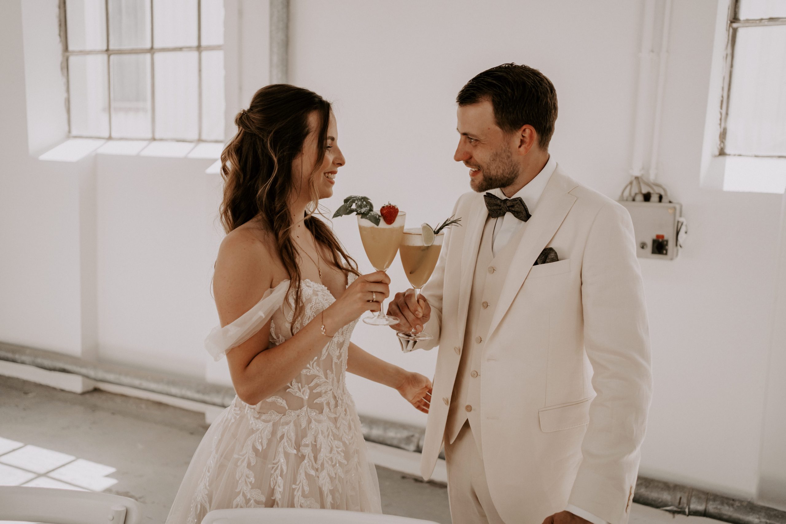 Bruid en bruidegom proosten met cocktails tijdens hun bruiloft, beiden gekleed in elegante witte trouwkleding, lachend en kijkend naar elkaar in een lichte, moderne ruimte.