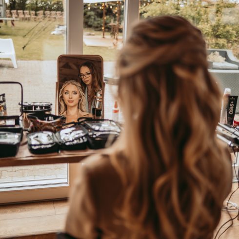 Achteraanzicht van een bruid die in de spiegel kijkt terwijl een visagiste haar kapsel en make-up perfectioneert voor de bruiloft, met een romantische buitenlocatie op de achtergrond.