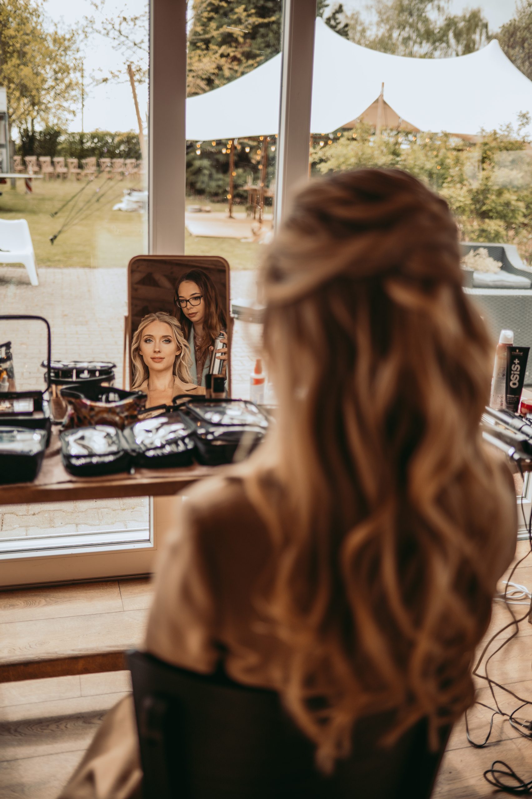 Achteraanzicht van een bruid die in de spiegel kijkt terwijl een visagiste haar kapsel en make-up perfectioneert voor de bruiloft, met een romantische buitenlocatie op de achtergrond.