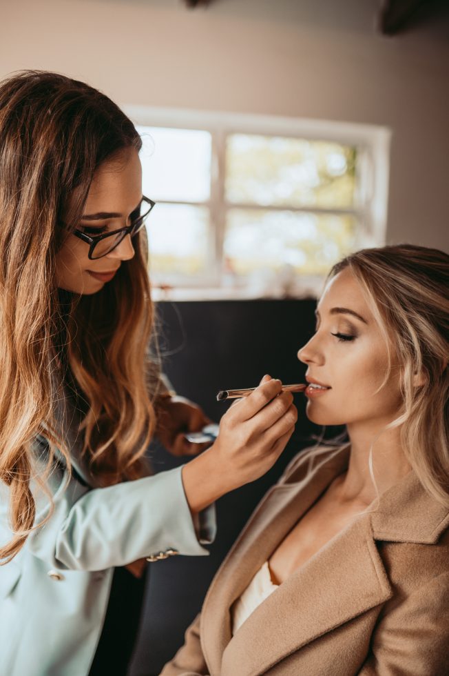 Close-up van een visagiste die lipmake-up aanbrengt bij een bruid met los opgestoken haar en een elegante beige jas, klaar voor de trouwdag.