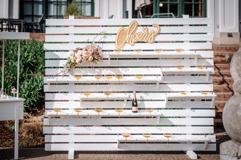 Witte houten prosecco- of champagnewand met het woord ‘Cheers’ bovenaan, gevuld met coupes met mousserende wijn, versierd met een bloemenarrangement en een fles champagne onderaan.