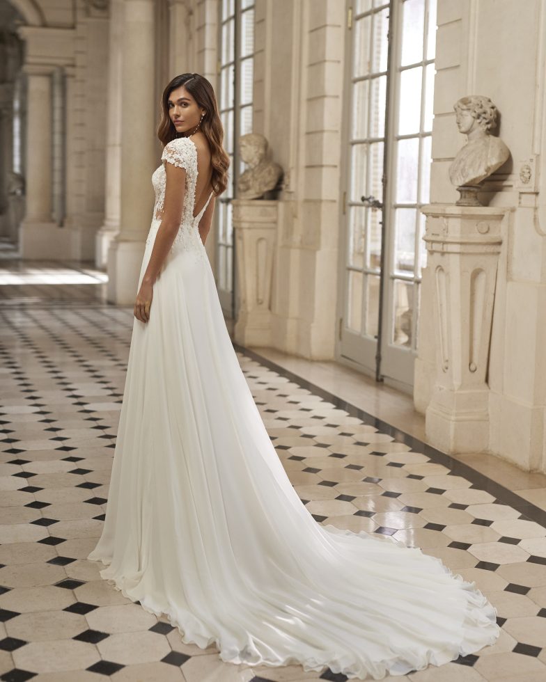 Bruid in een elegante trouwjurk met kanten lijfje en kapmouwtjes, staand in een klassieke, lichtovergoten galerij met zwart-wit geblokte vloer.