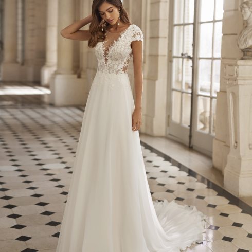 Bruid in een elegante trouwjurk met kanten lijfje en kapmouwtjes, staand in een klassieke, lichtovergoten galerij met zwart-wit geblokte vloer.