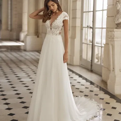 Bruid in een elegante trouwjurk met kanten lijfje en kapmouwtjes, staand in een klassieke, lichtovergoten galerij met zwart-wit geblokte vloer.