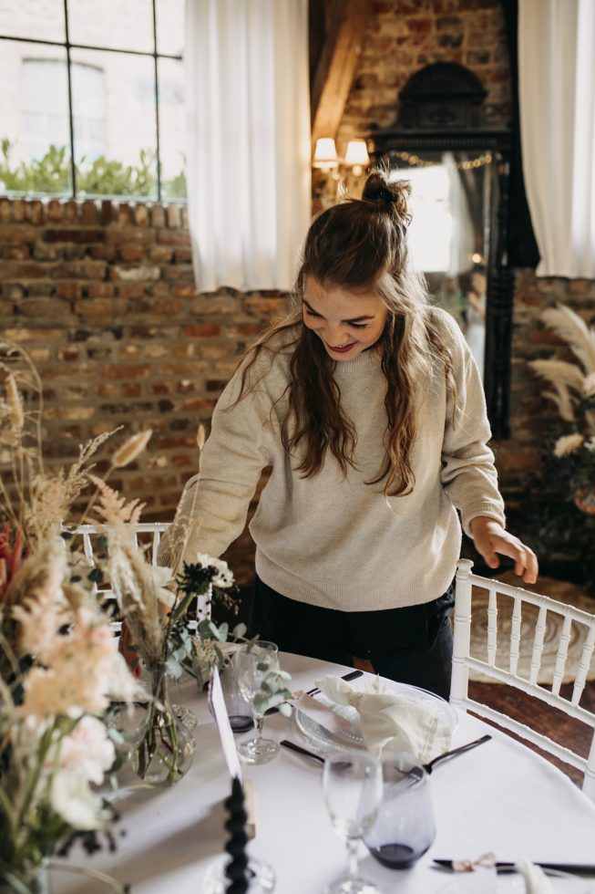 Vrouw met lang haar en een lichte trui dekt een feestelijk gedekte tafel met bloemen en servetten in een sfeervolle ruimte met bakstenen muur en grote ramen.