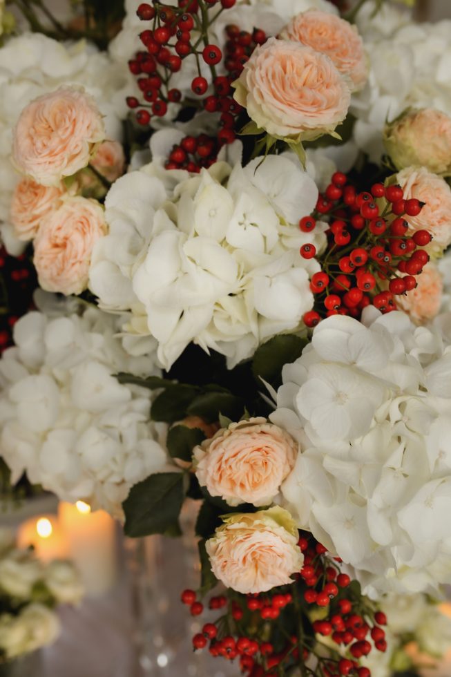 Elegante bloemencompositie met witte hortensia’s, zacht perzikkleurige rozen en rode bessen, met op de achtergrond zachte kaarsverlichting.