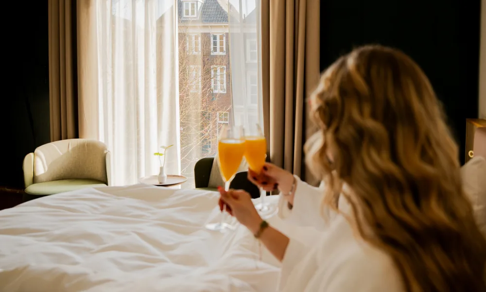 Vrouw in badjas geniet van ontbijt op bed met verse jus d’orange in een zonovergoten kamer bij Hotel Arsenaal in Delft, met uitzicht op de stad.