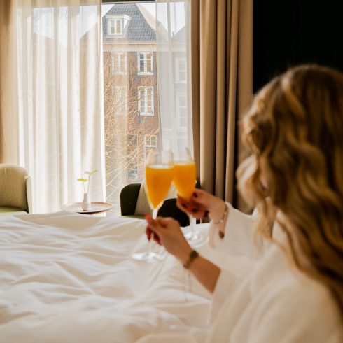 Vrouw in badjas geniet van ontbijt op bed met verse jus d’orange in een zonovergoten kamer bij Hotel Arsenaal in Delft, met uitzicht op de stad.