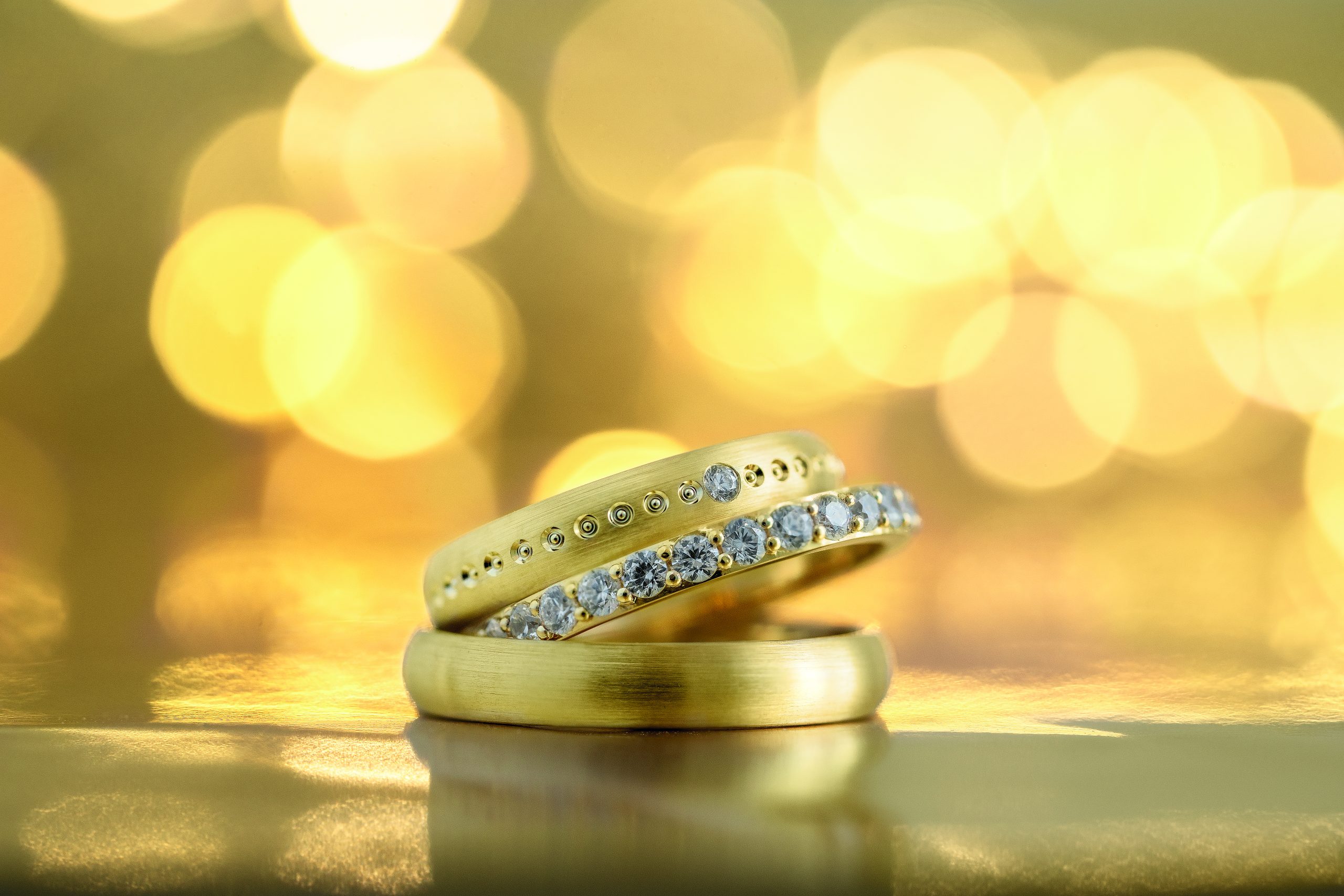 Close-up van drie gouden trouwringen met diamanten op een gouden achtergrond, perfect voor een luxe en stijlvolle bruiloft.