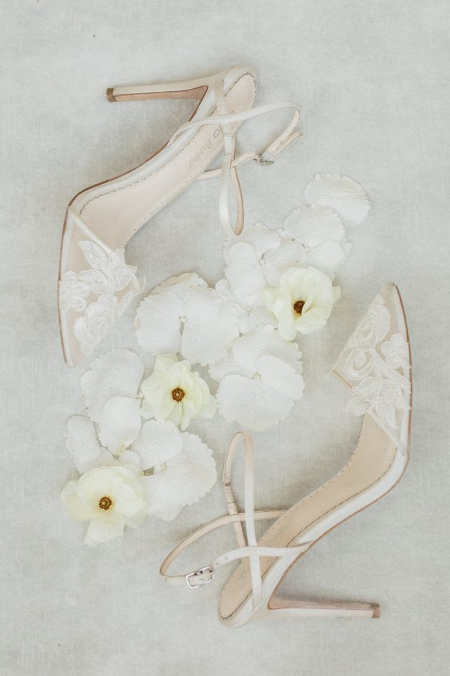 Elegante ivoorkleurige bruidsschoenen met fijne kanten details, artistiek gestyled met zachte witte en lichtgele bloemen op een lichte achtergrond.