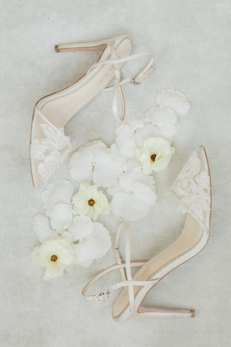 Elegante ivoorkleurige bruidsschoenen met fijne kanten details, artistiek gestyled met zachte witte en lichtgele bloemen op een lichte achtergrond.