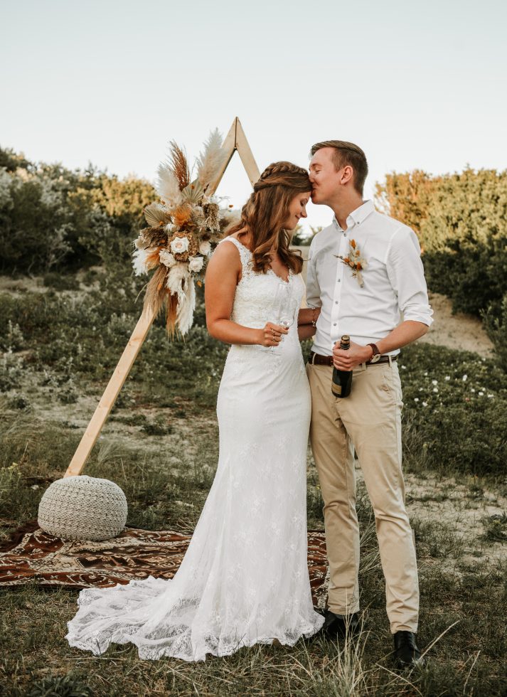 Bruidspaar staat liefdevol bij elkaar tijdens een intieme buitenbruiloft; de bruidegom geeft de bruid een zachte kus op haar voorhoofd terwijl ze beiden genieten van champagne, tegen een romantische achtergrond met een houten trouwboog versierd met bloemen en droogboeketten in natuurlijke tinten.