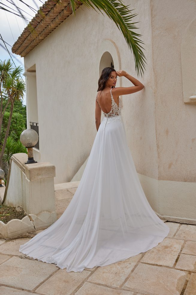 Bruid in een elegante trouwjurk met een kanten top en spaghettibandjes, poserend tegen een mediterrane gevel met zachte, natuurlijke tinten. Dit is de achterkant van de jurk