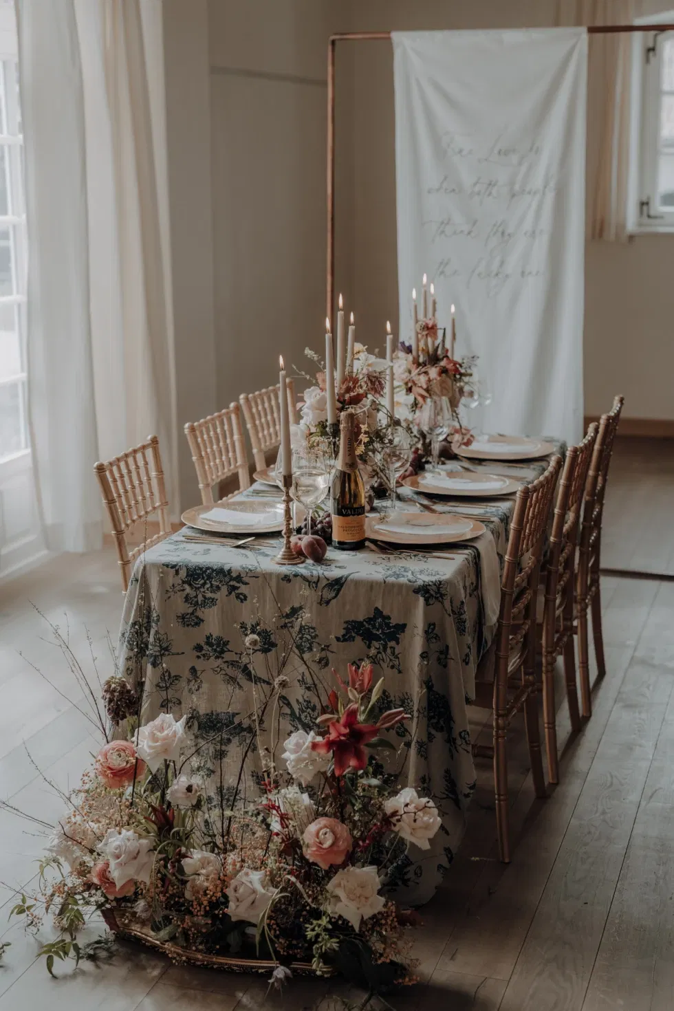 Stijlvol gedekte dinertafel met bloemenarrangementen, kaarslicht en een gebloemd tafelkleed, omringd door houten stoelen in een lichtovergoten ruimte.