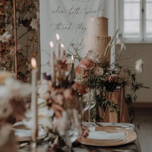 Bruiloftstafel met elegante bloemstukken, brandende kaarsen en een stijlvolle bruidstaart, tegen een backdrop met de tekst: 'True love is when both people think they are the lucky one.