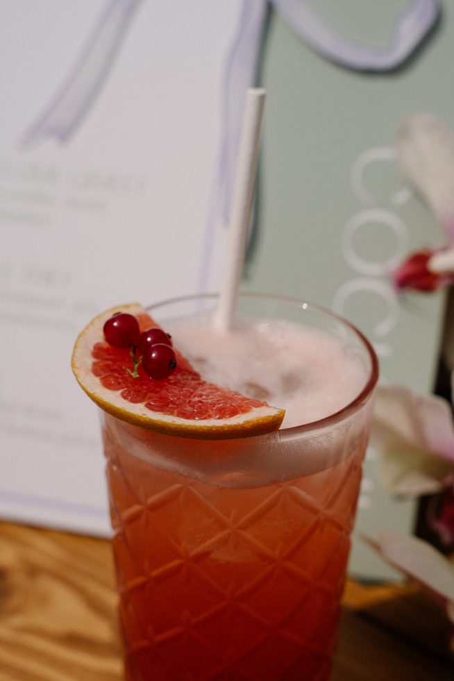 Close-up van een roze cocktail in een glas met ruitpatroon, gegarneerd met een schijfje grapefruit en rode bessen, met een rietje erin.