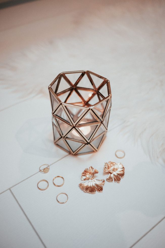 Stijlvolle gouden sieraden, waaronder ringen en opvallende oorbellen, liggen op een witte ondergrond naast een geometrisch windlicht met brandend kaarsje.